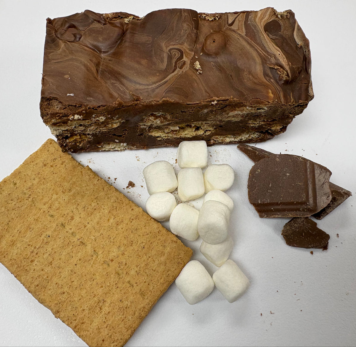 S'mores chocolate fudge with marshmallows, a graham cracker, and chocolate pieces arranged on a white surface.